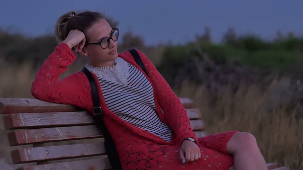 Dziewczyna w okularach siedzący na ławce, oglądając morze wieczorem - Materiał filmowy, wideo