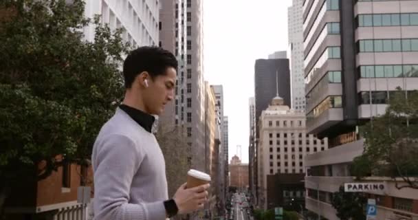 Side view of a young mixed race man walking in the city street with earphones on, holding a takeaway coffee cup with buildings in the background - Felvétel, videó