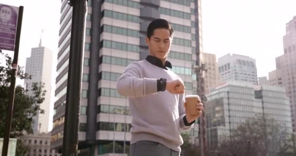 市街地に立つ若い混合レースマンのフロントビュー低角度テイクアウトコーヒーカップを保持し、背景の建物と時間をチェック - 映像、動画