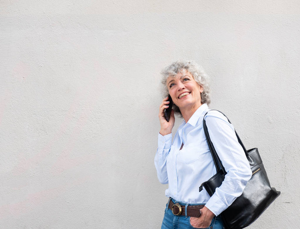 Side portrait of middle aged woman talking with mobile phone while leaning against white wall - Foto, afbeelding