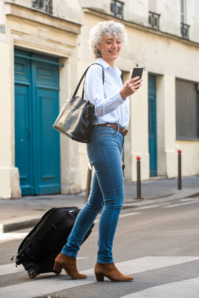 Retrato completo de mujer mayor feliz caminando con teléfono móvil y maleta en la calle
 - Foto, imagen