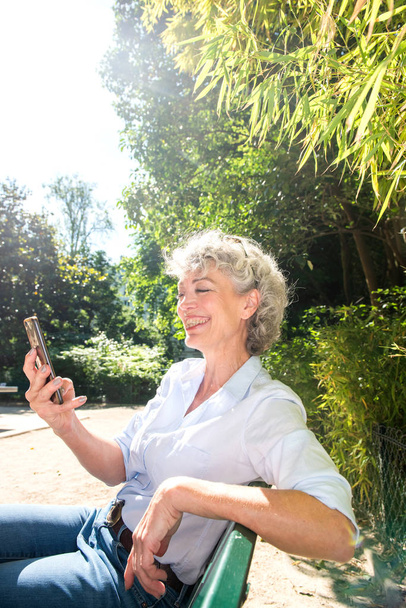 Zijportret van een oudere vrouw lachend en zittend in het park kijkend naar een mobiele telefoon  - Foto, afbeelding