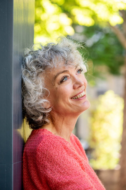 Retrato lateral de una atractiva mujer mayor apoyada en la pared sonriendo y mirando hacia otro lado en contemplación
 - Foto, Imagen