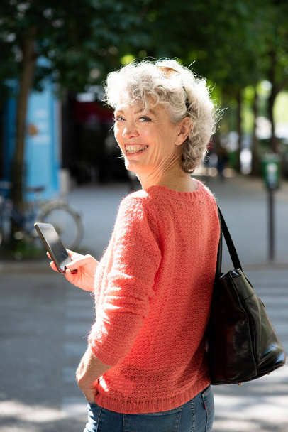 Portrait from behind of middle aged woman walking with mobile phone and bag in city - Photo, image