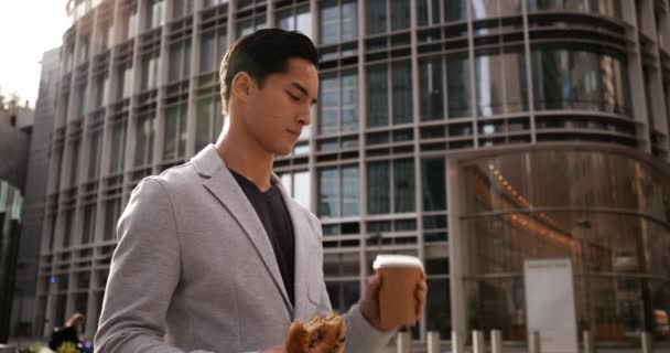 Front view close up of a young mixed race man standing, eating sandwich and drinking takeaway coffee during a lunch break in the city street with buildings in the background - Video, Çekim