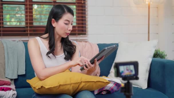 Moda asiática influencer diseñador mujeres utilizando la cámara de streaming y en vivo para vender ropa de negocios, el comercio electrónico transmitido en línea utilizando tableta chat respuesta cliente en el sofá en la sala de estar en casa. - Metraje, vídeo