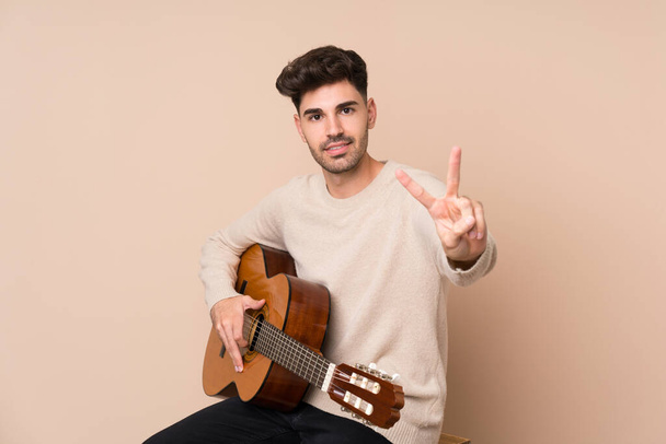 Young man with guitar over isolated background smiling and showing victory sign - Photo, image