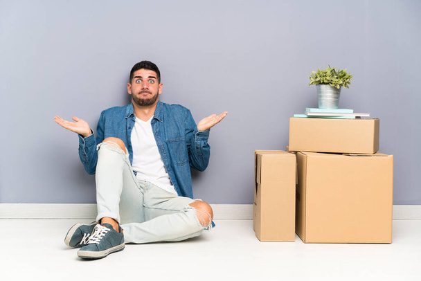 Handsome young man moving in new home among boxes having doubts with confuse face expression - Photo, Image