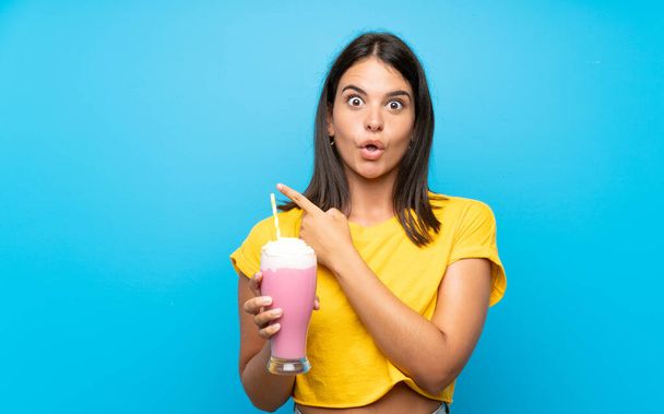 Молодая девушка с клубничным молочным коктейлем на изолированном фоне удивлена и указывает на сторону
 - Фото, изображение