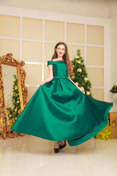 piękna wesoła smukła kobieta w długim zielonym sukni wieczorowej świętować Boże Narodzenie w urządzonym białym pokoju w pobliżu dużego złotego lustra. Korpus pełnej długości - Zdjęcie, obraz