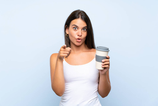 Mladá žena drží odnést kávu přes izolované modré pozadí překvapen a ukazuje dopředu - Fotografie, Obrázek
