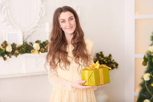kaunis hoikka valkoihoinen aikuinen nainen pitkä ilta keltainen mekko tilalla lahjapaketti keula ja juhlii joulua sisätiloissa hänen valkoinen huone lähellä takka
 - Valokuva, kuva