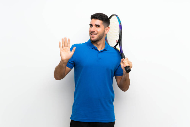 Przystojny młody tenisista człowiek nad odizolowaną białą ścianę salutując z ręki ze szczęśliwą ekspresją - Zdjęcie, obraz