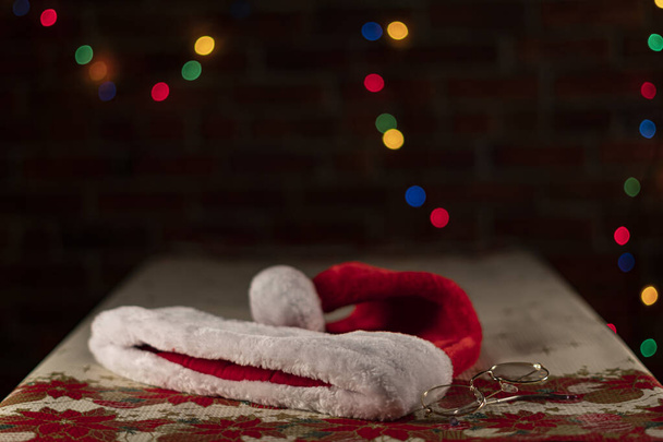 Weihnachtsmannmütze und -brille auf einem Weihnachtstisch mit unscharfem hellen Hintergrund an einer Ziegelwand. Weihnachtskonzept - Foto, Bild