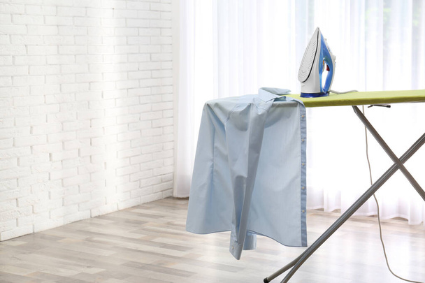 Board with modern iron and shirt at home. Laundry day - Φωτογραφία, εικόνα