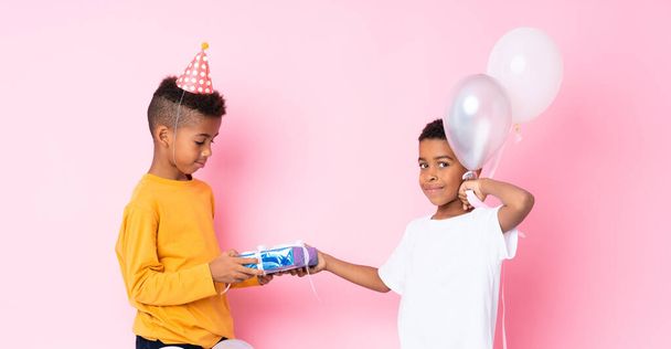 Dos hermanos afroamericanos sosteniendo globos y un regalo sobre un fondo rosa aislado
 - Foto, imagen