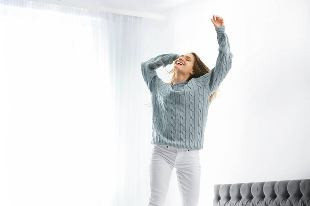 Young woman in warm sweater jumping on bed at home - Foto, afbeelding