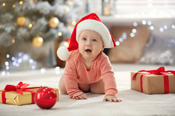 Little baby in Santa hat with Christmas decoration on floor at home - Fotoğraf, Görsel