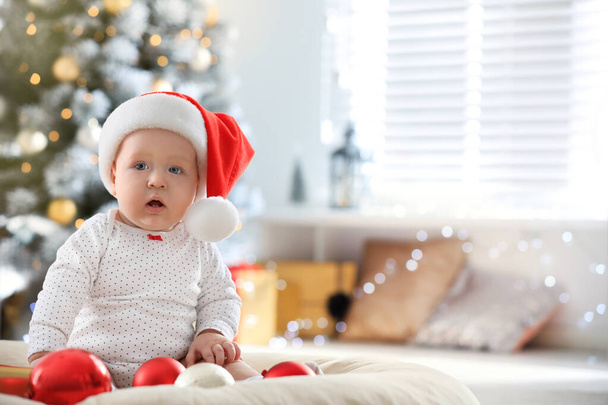 Little baby in Santa hat playing with Christmas balls indoors - Foto, Bild