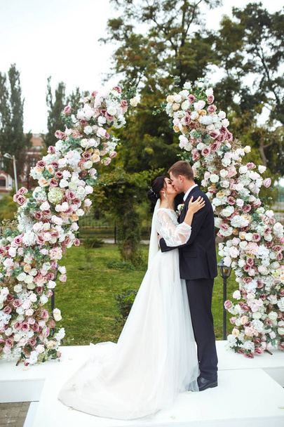 Loving newlyweds stand near the flower arch. Young beautiful happy wedding couple is kissing near arch at the ceremony. Pretty bride and stylish groom. Wedding and honeymoon concept. Love. - Foto, afbeelding
