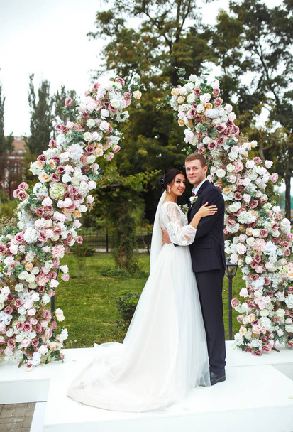 Loving newlyweds stand near the flower arch. Young beautiful happy wedding couple is kissing near arch at the ceremony. Pretty bride and stylish groom. Wedding and honeymoon concept. Love. - 写真・画像