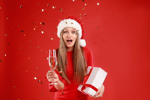 Emotional young woman in Santa hat with Christmas gift and glass of champagne on red background - Photo, image