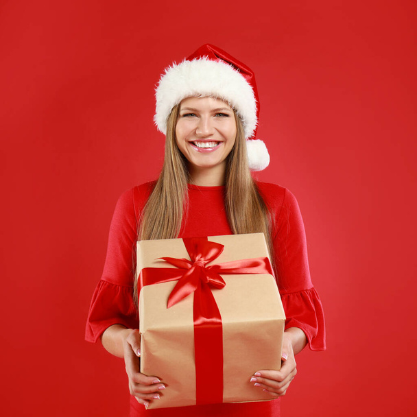 Happy young woman in Santa hat with Christmas gift on red background - Фото, зображення