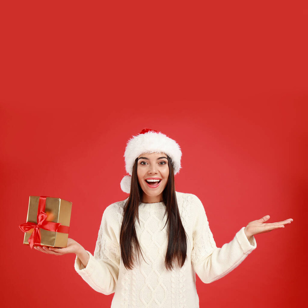 Happy young woman in Santa hat with Christmas gift on red background - Foto, afbeelding