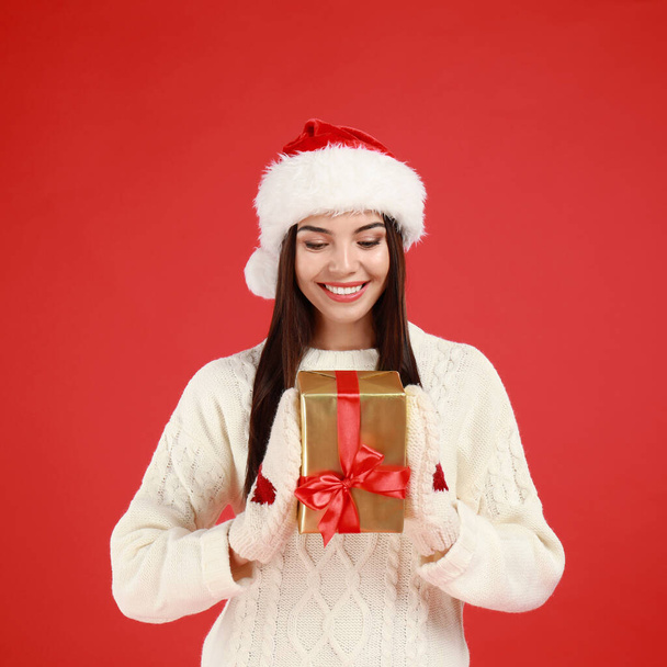 Happy young woman in Santa hat with Christmas gift on red background - Fotó, kép