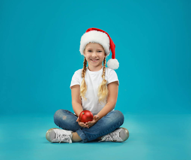 Cute little child wearing Santa hat holding Christmas decoration on blue background - Photo, image