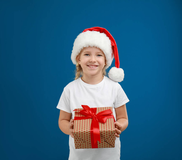Cute little child wearing Santa hat with Christmas gift on blue background - Фото, изображение