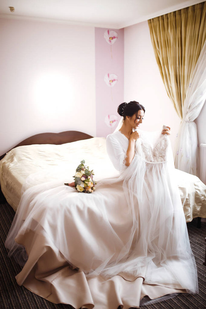Morning of beautiful bride near the wedding dress. Bride in a white robe  holds and  admires her wedding dress. Beautiful woman with professional make up and hair style. Morning of the bride.  - Photo, image