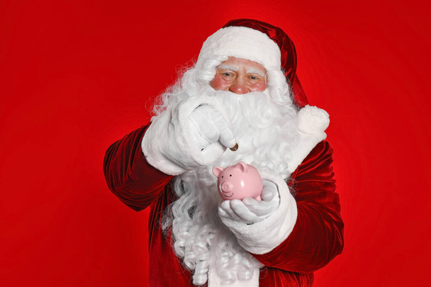 Santa Claus putting coin into piggy bank on red background - Fotó, kép