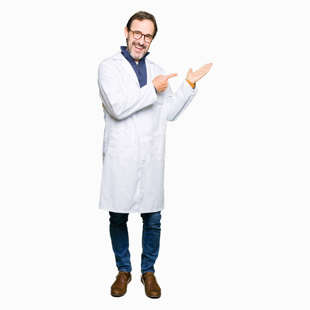 Middle age therapist wearing white coat amazed and smiling to the camera while presenting with hand and pointing with finger. - Photo, Image