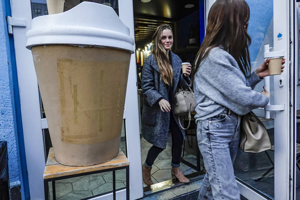 Kiev, Ucraina Due giovani donne con tazze di caffè sono giustapposte con una tazza di caffè di carta di grandi dimensioni fuori da un caffè nella sezione Podil della città
. - Foto, immagini