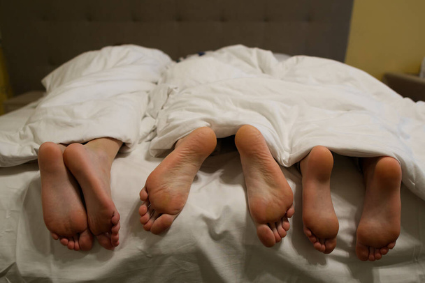 τρία ζευγάρια πόδια στο κρεβάτι κάτω από μια λευκή κουβέρτα - Φωτογραφία, εικόνα