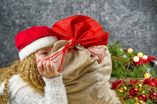 Malá blondýnka dívka v gnómové červené klobouk s sáčkem santas v očekávání vánočního zázraku. - Fotografie, Obrázek