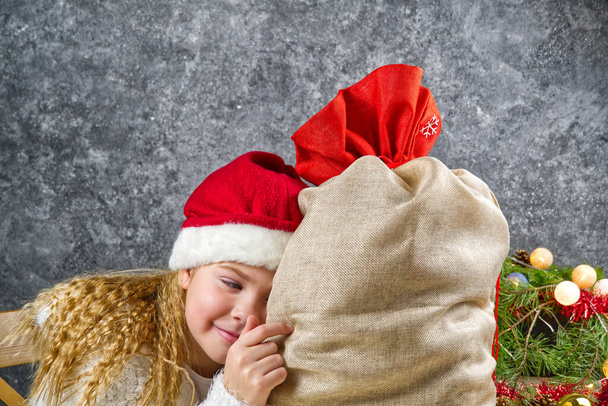 Niña rubia en un sombrero rojo gnomos con bolsa de santas en anticipación de un milagro de Navidad
. - Foto, Imagen