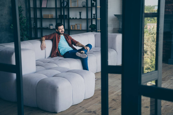 Photo of handsome guy in living room sitting comfortable couch relax saturday evening holding leg on knee modern apartment indoors casual dressed - Foto, Imagem