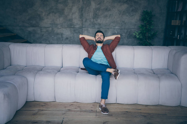 Full length photo of handsome guy in high spirits holding hands behind head sitting couch sleeping relaxing saturday morning in modern flat loft indoors - Foto, immagini