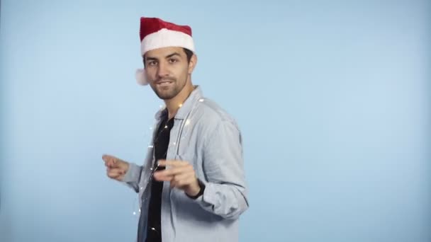 Positive handsome man in a Christmas red hat and garland on neck dances and smiles at the camera - blue wall background studio. Man in casual clothes and hat freerly dances in front the camera - Video