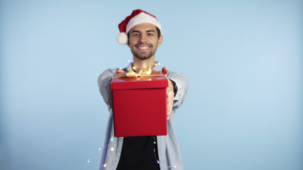 Young man wearing Christmas Santa hat and garland on neck holding gift over isolated blue background with outstretched hands with a happy face standing and smiling with a confident smile showing teeth - Filmagem, Vídeo