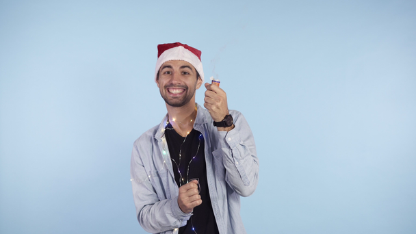 Young, smiling man in casual clothes and santa hat on a head exploding confetti cracker on a blue background. Happy celebrating of a New Year or Chrismas - Video