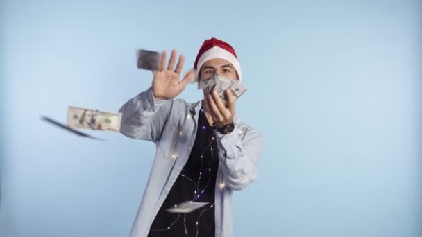 Crazy, handsome excited man throwing dollar banknotes and smiling. Squandering, wealth. Man in a santa red hat and garland on neck standing isolated on blue background - Video, Çekim