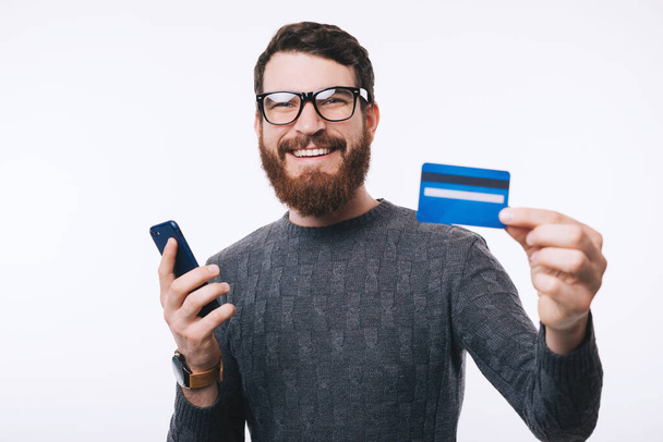 Ευτυχισμένος όμορφος άντρας με γυαλιά που δείχνει στη νέα του πιστωτική κάρτα ένα κινητό τηλέφωνο. - Φωτογραφία, εικόνα