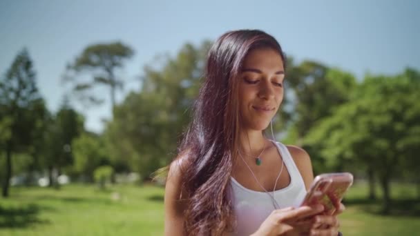 Portrait of a happy brunette young woman standing in the park with earphones in her ears texting message on smartphone - young college student on her phone - Materiaali, video