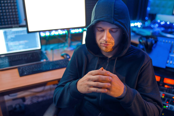 Hacker mit Maske und Kapuze sitzen an seinem Arbeitsplatz mit Laptop und PC und hacken Informationen. Internet-Spionage, illegaler Lebensstil - Foto, Bild