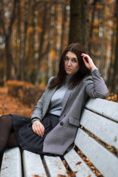 Herbst im Freien Porträt einer jungen Frau im Herbst Park, sitzt auf einer weißen Bank und genießt die Herbstlandschaft, trägt graue Jacke, graues Badlon und schwarzen Rock. Bunte gefallene Blätter auf dem Hintergrund - Foto, Bild