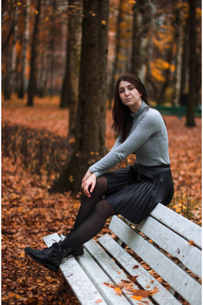 Podzimní portrét mladé ženy v podzimním parku, sedící na zadní straně bílé lavičky a užívající si podzimní krajiny, oblečená v badlonu a černé sukni. Barevné spadlé listí a stromy na pozadí - Fotografie, Obrázek