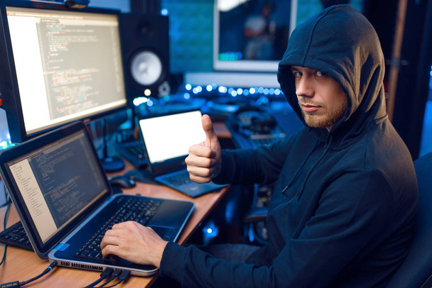 Hacker in hood toont duimen op zijn werkplek met laptop en PC, wachtwoord of account hacking. Internet spion, criminaliteit levensstijl, risico baan, netwerk crimineel - Foto, afbeelding